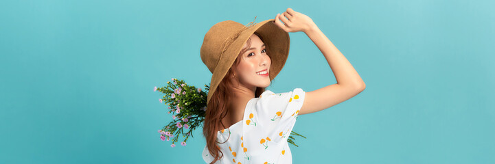 Image of happy woman in elegant dress and straw hat holding flowers isolated over blue background