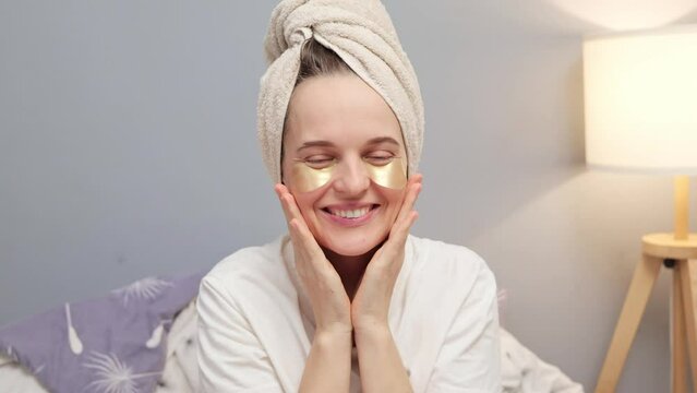 Funny positive woman wrapped in towel sitting in bedroom with under eyes patches sending air kissing while enjoying skin care procedures beauty treatment routine