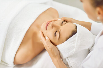 Happy beautiful young woman with white towel turban on head relaxing and enjoying facial treatment...