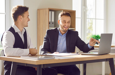 Two happy smiling young coworkers and company employees sitting at the desk on workplace on meeting looking at laptop monitor screen and discussing new projects or startups, analyzing company.