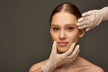 estheticians in medical gloves examining face of woman on grey background, dermatology concept