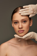 esthetician in medical gloves touching face of young woman on grey background, dermatology concept