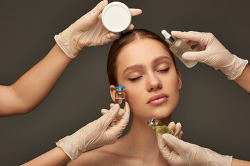 estheticians in medical gloves holding beauty treatment products near woman on grey backdrop
