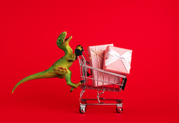 Funny green dinosaur toy with shopping cart  with present boxes on red background. Holiday shopping concept.