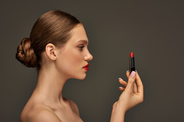side view of beautiful young woman holding red lipstick on grey background, beauty trends