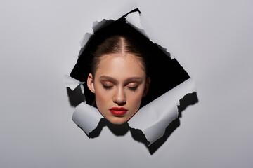 pretty young woman with blue eyes and red lipstick peeking through hole in ripped grey paper