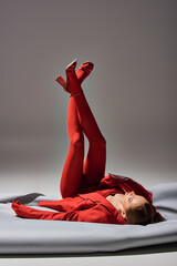 full length of stylish young model in red outfit posing with raised legs in shoes on grey background