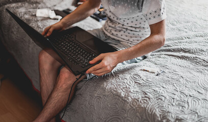 A young man with a laptop on his knees is sitting on the bed.