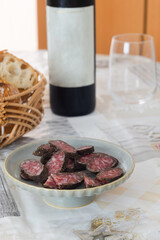 A plate of sliced pure pork dried sausage with crust black peppers and bottle of wine and basket of bread in background 