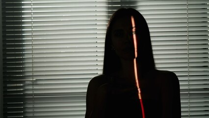 Portrait of female model in studio. Appealing woman silhouette with light pointing on part of the body and face standing against window with jalousie.