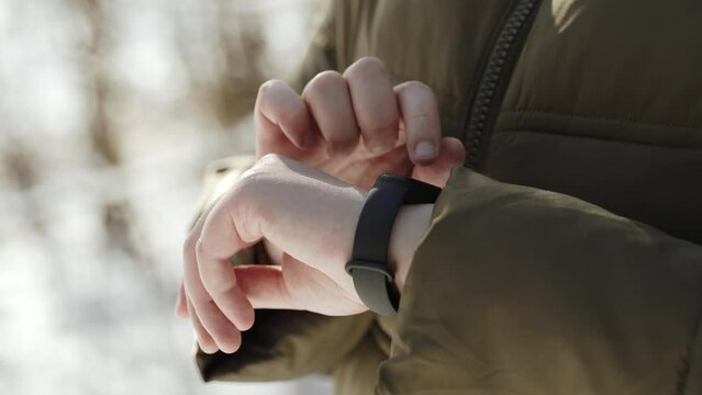 Woman in warm clothes taps finger on touch fitness bracelet in sunny winter. Unrecognizable lady looks at notifications on her smart watch and health indicators during frosty and snowy walk.
