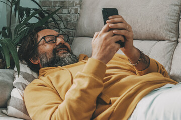 One mature man use mobile phone cellular laying on tne sofa at home in relax leisure online activity. People using cell smartphone indoor in apartment. Concept of real life and surf the web alone - obrazy, fototapety, plakaty