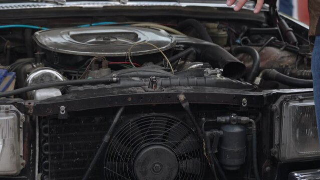 Man points with hand at carburetor engine of old car with cooling radiator outdoor. Breakdown of car standing with hood raised while traveling. Cooling system with fan and headlights of sedan.