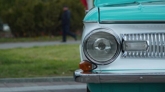 Front round one headlight and false grille of vintage white turquoise car in parking lot on cloudy day. Unrecognizable man in black clothes is walking along path in background. Details of old car.