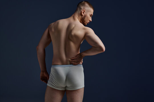 Back view portrait of young man with perfect body shapes in underwear put his hand on back as he have pain against blue background. Concept of men's health, lifestyle, beauty, body and skin care