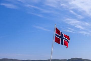 A vibrant Norwegian flag unfurling against a cerulean sky on a sunlit summer day, with the lower part of the image showcasing a distant range of gentle northern mountains