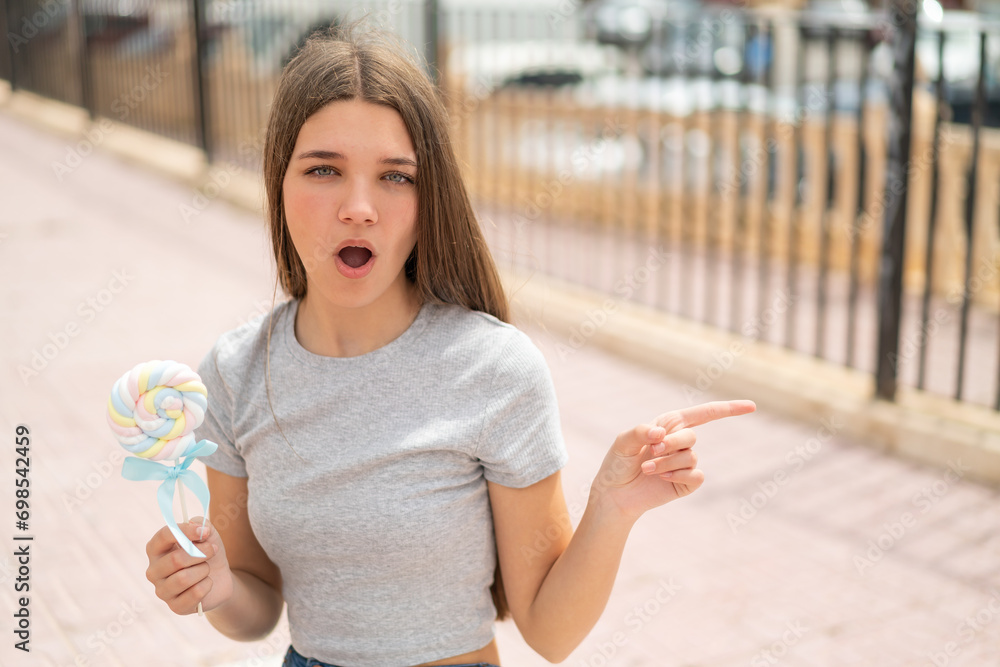 Poster Teenager girl holding a lollipop surprised and pointing side