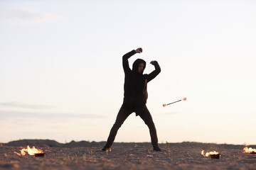Full length portrait of young man twirling fire rod during outdoor fire show performance on beach,...