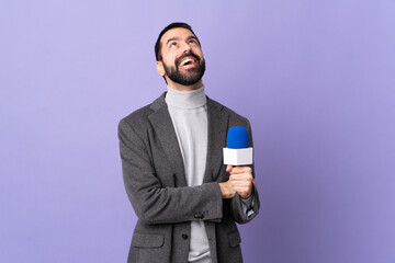 Adult reporter man with beard holding a microphone over isolated purple background looking up while...