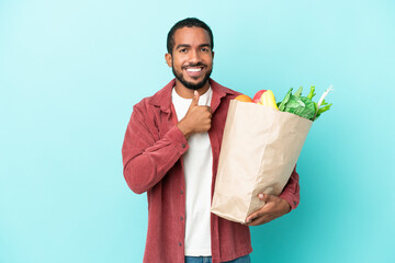 Young latin man holding a grocery shopping bag isolated on blue background giving a thumbs up...