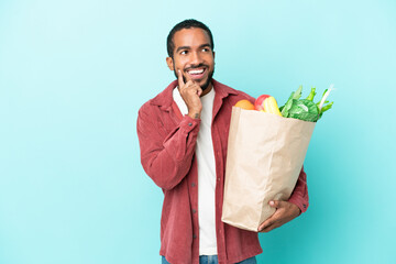 Young latin man holding a grocery shopping bag isolated on blue background thinking an idea while...