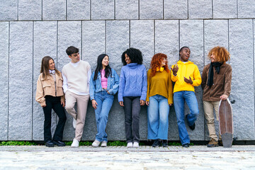 Diverse group of seven young friends enjoying time together by a gray wall, showcasing racial...