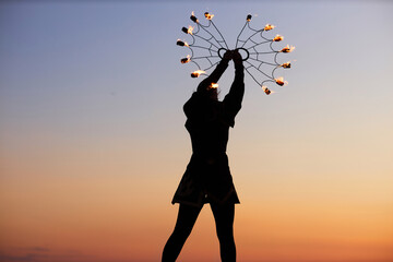 Silhouette of young woman dancing with fire fans against sunset sky in nature, copy space