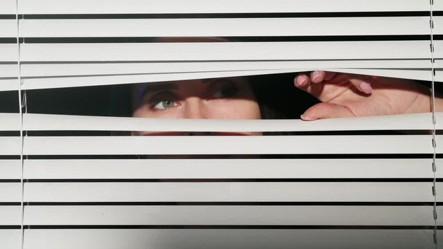Portrait of attractive female model in studio. Close up shot of the window with white blinds, brunette woman opens part of it and looking out.