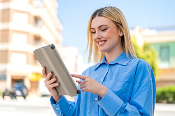 Young pretty blonde woman at outdoors touching the tablet screen with happy expression