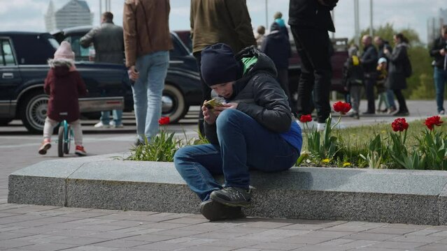 Lonely lost boy tries to contact his parents via smartphone. Sad child sits on parapet of flowerbed in the city near vintage car parking lot. People pass by indifferently.