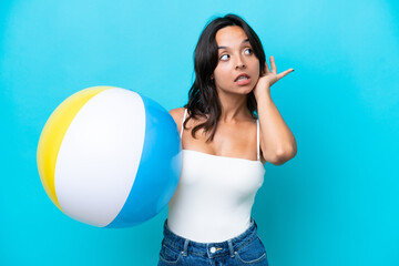 Young hispanic woman holding beach ball isolated on blue background listening to something by putting hand on the ear