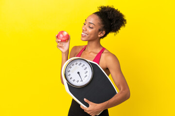 Young African American woman isolated on yellow background with weighing machine and with an apple
