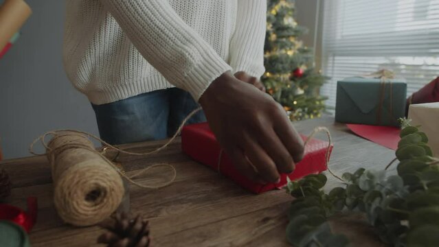 Cropped shot of black woman using twine while wrapping Xmas gift in red paper crafting on table