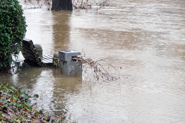 Konzept Hochwasser: Blick auf ein überflutetes Ufer im Dauerregen mit einer Bank und Mülleimer...