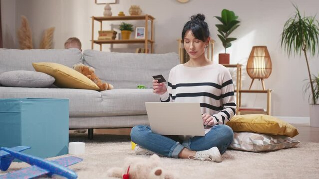 Woman adding private details into laptop. Female entering card number into laptop. Woman sitting at floor carpet and working with laptop. Female holding bank card and smiling at camera.