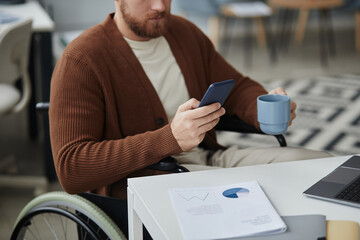 Close up of young man with disability working at desk in office and using smartphone during coffee...