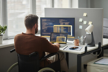 Back view of adult man with disability as IT professional using computer and writing code at...
