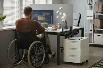 Back view of adult man with disability as computer programmer writing code at workplace in office,...