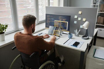 Back view of adult man with disability using wheelchair at office workplace and coding in IT development - obrazy, fototapety, plakaty