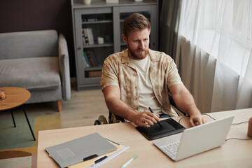 High angle portrait of young bearded man with disability using pen tablet while working as digital artist at home office, copy space