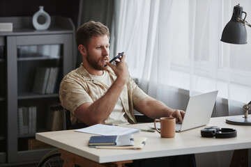 Portrait of bearded adult man with disability working at home office workplace and recording voice message via smartphone, copy space