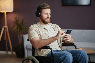 Portrait of bearded man with disability wearing headphones at home and listening to music via smartphone, copy space - obrazy, fototapety, plakaty