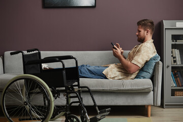 Side view portrait of man with disability relaxing on couch at home and using smartphone with wheelchair in foreground, copy space