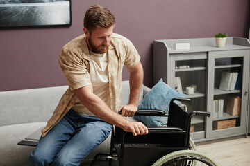 Portrait of independent adult man with disability getting into wheelchair in home setting, copy space