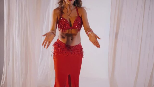 Beautiful girl dances oriental belly dance. Sexy woman in red lingerie is dancing a seductive dance in a white studio. Slow motion shooting. Beautiful stomach and breasts.