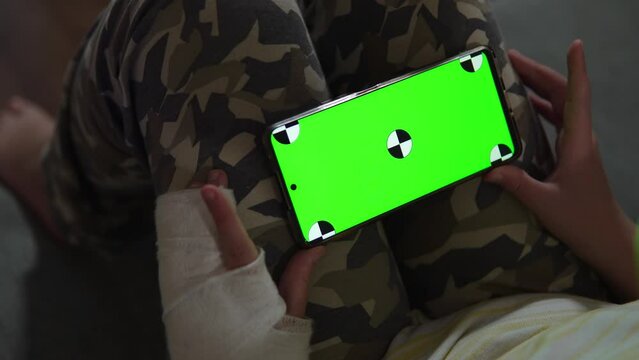 Child with cast on his arm looks at phone screen on knees at home, view over the shoulder. Green phone screen with chromakey marks. Entertainment during temporary physical limitations.