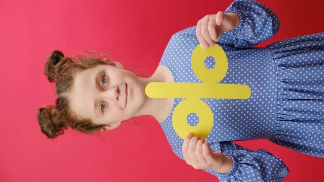 Vertical footage of pleasant happy little girl holding yellow percent mark, expressing positive emotions, presenting discounts, isolated on red background wall in studio. Sale concept. Selective focus