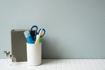 Notebook and holder with glue stick, pen, scissors on white tile desk. gray wall background