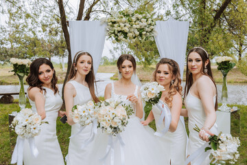 Group portrait of the bride and bridesmaids. Bride in a wedding dress and bridesmaids in white dresses and holding stylish bouquets on the wedding day against the background of the lake.