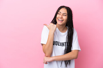 Teenager volunteer girl with braids isolated on pink background pointing to the side to present a...